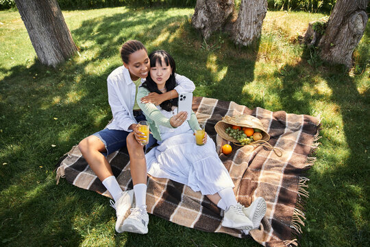 Two young women, one African American and one Asian, enjoy a picnic outdoors while taking a selfie.