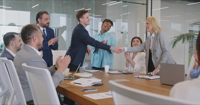 Two diverse teams meet and make a business deal. Happy man and woman shake hands over the negotiation table while employees applaud. Professional partnership, cooperation, teamwork, success concept