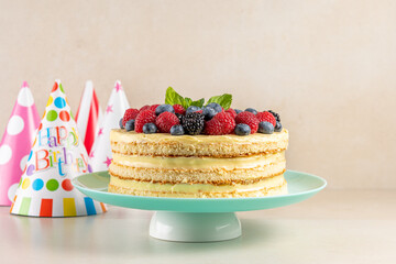 Homemade cake with fresh berries and birthday hat on bright background.