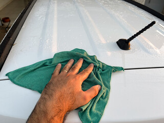 A man's hand uses a green microfiber cloth to wipe down a white car.