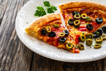 Circle pizza chorizo slices with black and green olives and mozzarella served on white plate on wooden table 