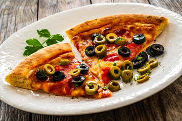 Circle pizza chorizo slices with black and green olives and mozzarella served on white plate on wooden table 