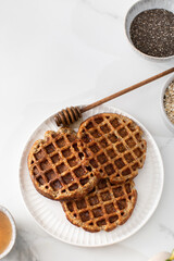 Homemade oatmeal and chia seed waffles on a white plate, accompanied by honey and ingredients
