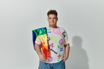 A queer person with a rainbow flag draped over their shoulder, wearing a colorful t-shirt.