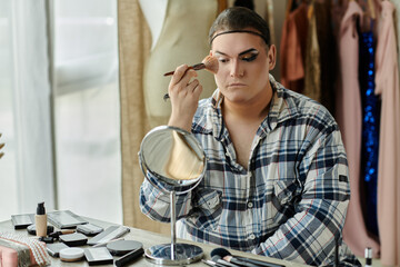 A queer person applies makeup in front of a mirror, surrounded by various cosmetic products.