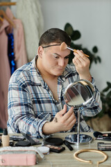 A queer person applies makeup while looking in a mirror, surrounded by beauty products.