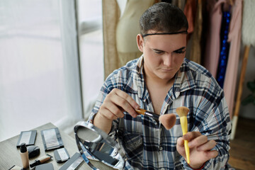 A queer person applies makeup while looking in a mirror, surrounded by a collection of beauty products.