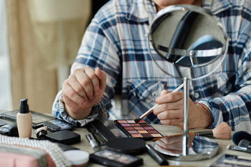 A queer person applies eyeshadow while looking in a mirror, surrounded by makeup products.