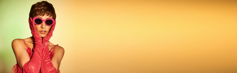 Fashionable young woman in pink attire and sunglasses posing elegantly on vibrant backdrop.