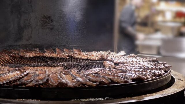 Process of Cooking Juicy Smoked Pork Ribs on a Large Rotating Charcoal Grill. Close up. Chef preparing fried ribs on a flaming grill or brazier in a restaurant. Pork ribs cooked on fire in smokehouse.