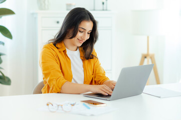 Confident successful amazing brunette arabian or indian ethnicity young woman, IT specialist, product manager sits at a workplace working in a laptop on a project, looking device screen, smiles