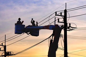 Silhouette of 2 lineman electricians on bucket boom truck are repairing electrical system on electric power pole against sunset sky background in evening time © Prapat