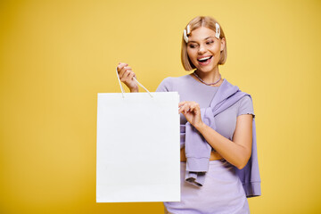 joyful attractive woman with accessories in chic attire posing with shopping bag on yellow backdrop