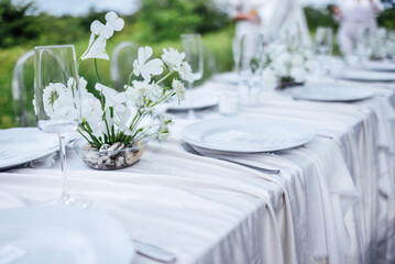 The festive table with a white tablecloth is decorated with flowers. Empty plates and glasses waiting for the guests. A holiday lunch in the garden.