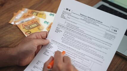 Businessman reviews Employees Withholding Certificate form with pen sitting at wooden surface with euro banknotes and laptop computer placed in front of man slow motion