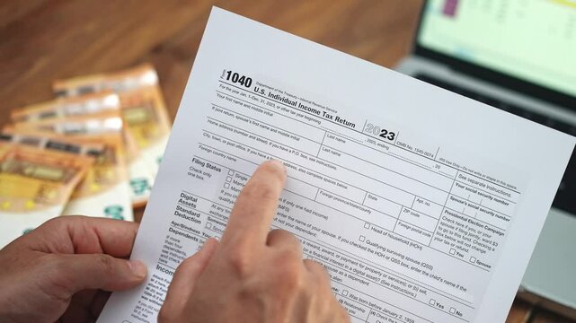 Male accountant examines 1040 tax form with finger highlighting details, man sits at wooden desk with prepared euro banknotes and laptop nearby slow motion