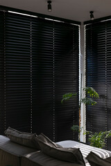 Wooden electric blinds in the modern interior, creating a sleek and stylish atmosphere. Sunlight filters through the venetian blinds on windows, black color, shadows on the sofa. Motorized shutters.