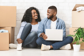 Moving concept. Happy african couple purchasing furniture for new apartment, sitting on floor and using laptop, free space