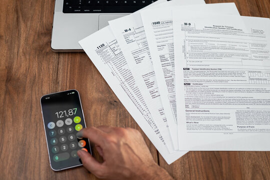 Man using a calculator and reviewing tax forms on a wooden desk. Financial planning and tax preparation concept. Essential for accountants, tax season, and personal finance management 