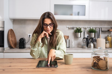 Smiling young woman wearing glasses drinking coffee and using tablet computer while sitting in kitchen. Businesswoman reading emails at morning in her home. Remote work business female