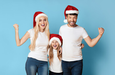 Obraz premium A family of three, wearing Santa hats and white t-shirts, pose with their arms raised in the air and smiling excitedly in front of a blue background.