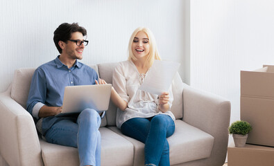 Obraz premium A young couple sits on a couch, discussing home improvement plans. The woman holds paperwork while the man uses a laptop.