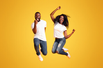 We made it. Joyful Afro Couple Jumping Up And Down, guy clenching his fist, yellow studio background