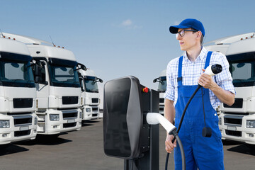 Driver in uniform holds electric vehicle charging plug. Electric trucks in the background