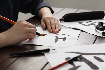 Naklejka premium Calligraphy. Woman with brush and inkwell practicing writing Chinese hieroglyphs on paper at wooden table, closeup