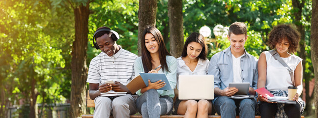 Education concept. Group of college students with laptop and tablet computer studying together outdoors in campus, sitting on bench outside