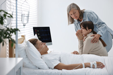 Coma patient. Sad women near unconscious young man in hospital