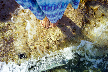 A woman in a dress enters the water: a photo of a woman's legs standing among clear water on the surface of a rock. Summer vibes in Montenegro: cooling the body in the sea.