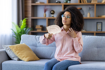 Woman sitting on couch in living room using hand fan to cool down. Expressions of discomfort due to...