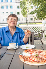 An elderly woman sits in a summer cafe, drinks coffee and pizza.