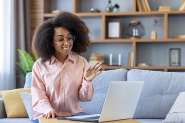 Smiling woman with curly hair participates in a video call, waving at the screen while sitting on a comfortable couch with laptop in modern living room