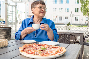 An elderly woman sits in a summer cafe, drinks coffee and pizza.