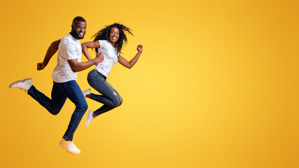 A couple, both wearing white t-shirts and jeans, leaps in the air with joyful expressions, against a vibrant yellow background.