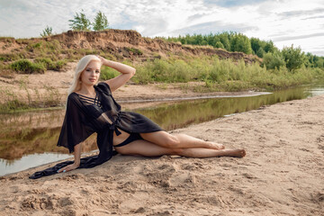 A girl sits on a sandy beach in a black swimsuit and cape.