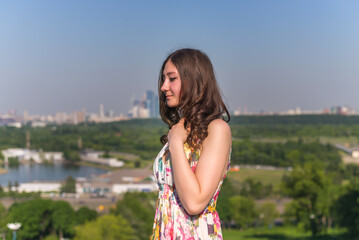 Fototapeta premium A young happy girl in a bright dress standing on a green grassy hillside on a sunny day enjoying the view of the distant city.