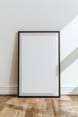 A blank white picture frame with a black border resting against a white wall on a wooden floor.