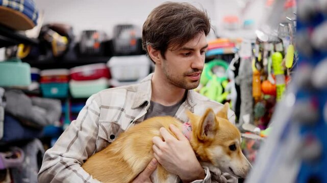 A confident brunette guy in a white shirt holds a red corgi dog in his hands and chooses food for it in a pet store