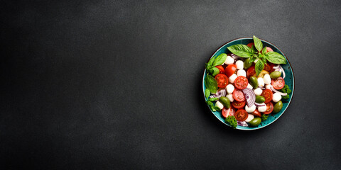 Caprese salad in a bowl: tomatoes, mozzarella cheese, basil and olives. On a black stone canvas. Minimalism style. Top view.