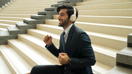 Project manager listening and enjoy music from headphone while sitting at stairs. Professional businessman wearing suit outfit while dancing and moving to music. Happy man listen funny song. Exultant.