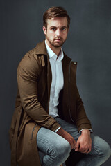 Fototapeta premium Style, portrait and man on chair in studio for confidence, trendy fashion or classy clothing on dark background. Smile, jeans and model on stool for elegant outfit, designer coat or semi formal