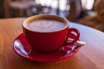 Close-up view of red porcelain cup of cappuccino with heart shaped froth art stands on wooden table in sidewalk cafe or street restaurant. Soft focus. Hot drinks theme.
