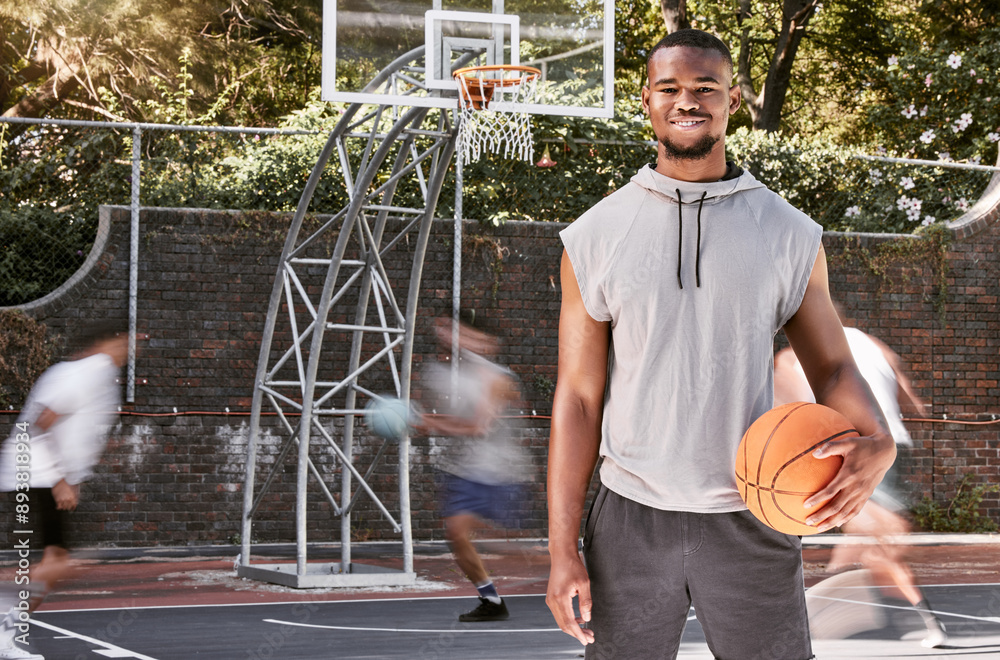 Sticker Portrait african american man standing with a basket ball on the court. Handsome male basketball player holding a sports ball while his friends and teammates play and practice in the background