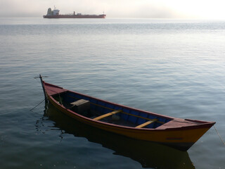 a small boat in the foreground on calm water with a larger ship and misty horizon in the background. It’s interesting due to the contrast in vessel sizes and the serene setting