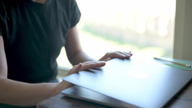 A person in a black shirt closing a laptop at a wooden desk by a bright window. The image captures the end of a work or study session in a modern, sunlit environment. High quality 4k footage