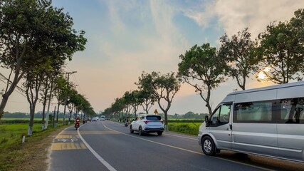 A quiet, scenic road lined with trees, cars, and motorcycles during sunset, highlighting rural transportation and peaceful travel