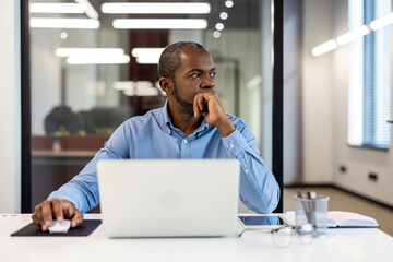 Professional businessman sitting at office desk with laptop, deep in thought. Modern workspace with...